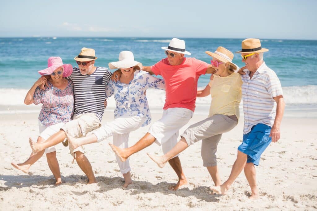 Group of senior friends laughing at the beach, arms around each other, kicking in unison