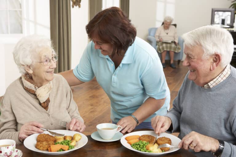 Two seniors eating at table, smiling woman greeting them