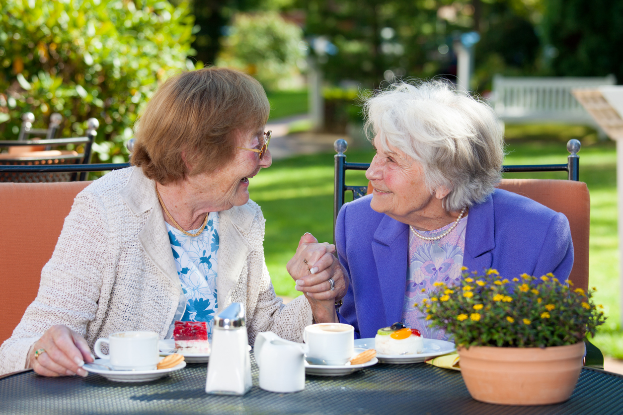 Two Happy Elderly Women Chatting at the Garden Table with Coffee and Snacks While Holding their Hands.