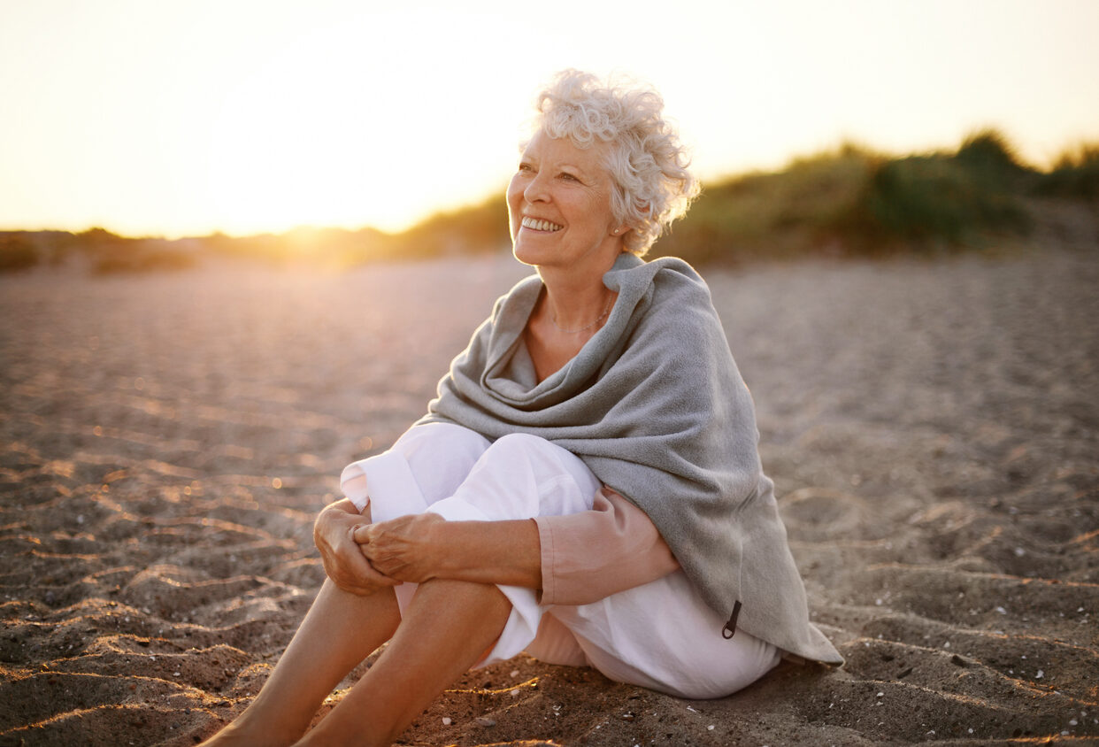 Happy retired woman wearing shawl sitting relaxed on sand at the beach. Senior caucasian woman sitting on the beach outdoors