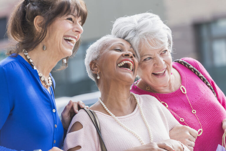 A group of three multi-ethnic senior women in their 60s and 70s standing outdoors, laughing. They are carrying shopping bags.